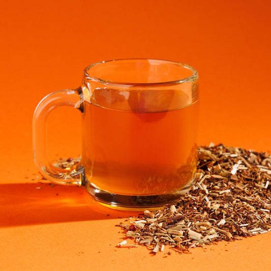 Glass cup of orange Evensong Chai tea positioned next to a pile of loose herbals on an orange background. A Good Store product.