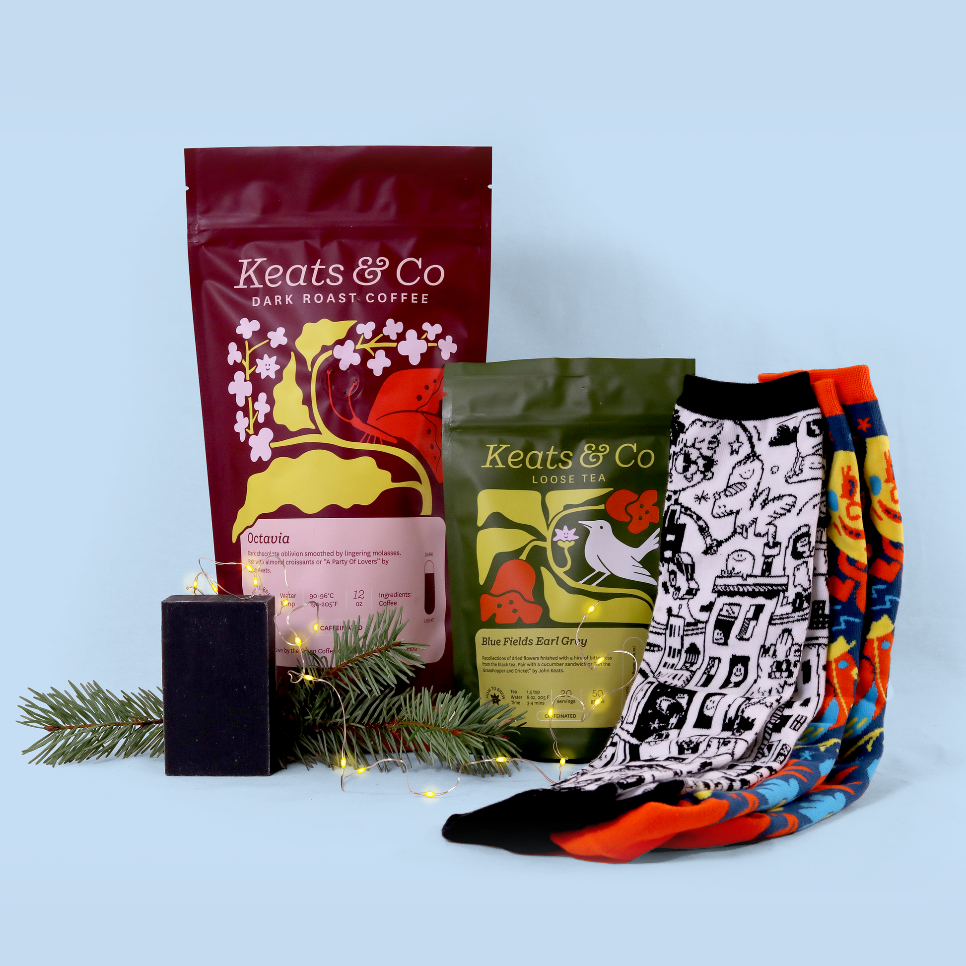 A product display of Keats & Co merchandise against a light blue background. The arrangement includes a burgundy bag of 'Octavia' dark roast coffee, a green package of 'Blue Fields Earl Grey' loose tea, a black candle, decorative socks in two patterns (one black and white with an artistic design, one with bright orange and blue colors), and a pine branch with small LED lights.