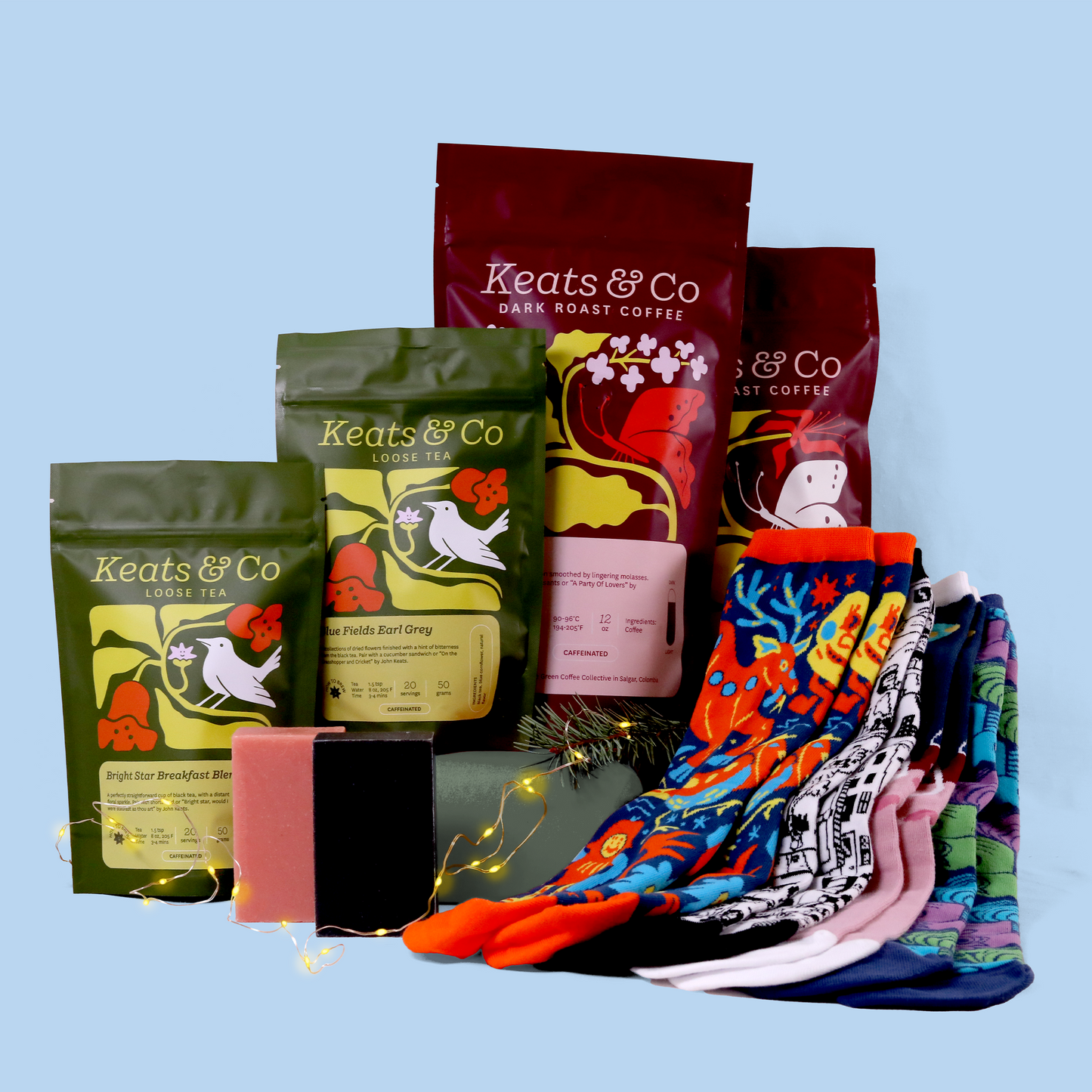 A product display of Good Store products against a light blue background. The arrangement includes multiple packages: two burgundy bags of dark roast coffee, two green bags of loose tea (Blue Fields Earl Grey and Bright Star Breakfast Blend), black and pink soap bars, and a collection of colorful patterned socks fanned out on the right side. Small LED lights are woven through the display, adding a decorative touch. 