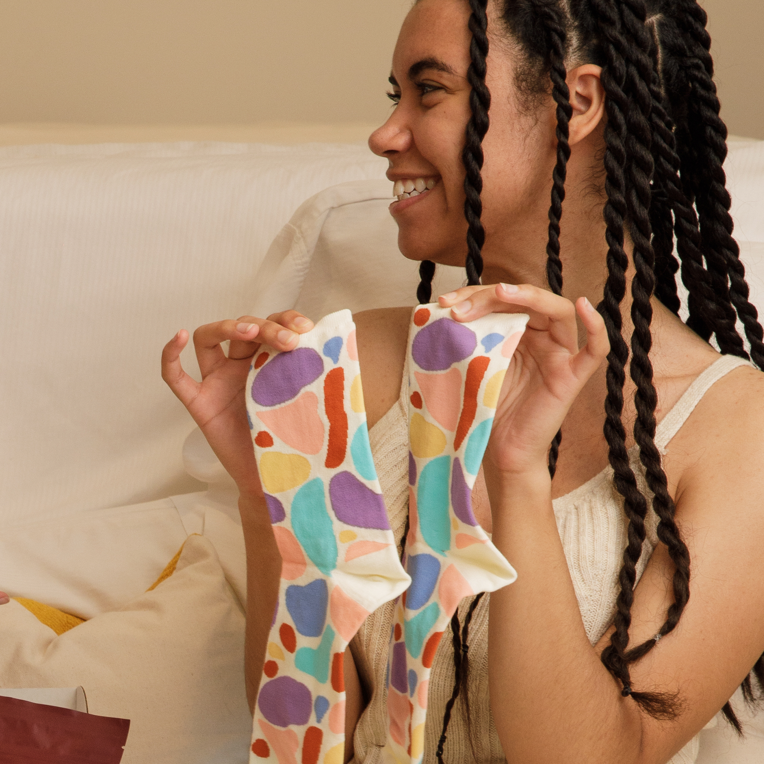 lose-up shot of a person with long black braids smiling while holding up colorful patterned Awesome Socks. They're wearing a white knit top and sitting on a light-colored couch. The socks feature an abstract pattern with purple, turquoise, pink, yellow, and orange shapes on a white background.