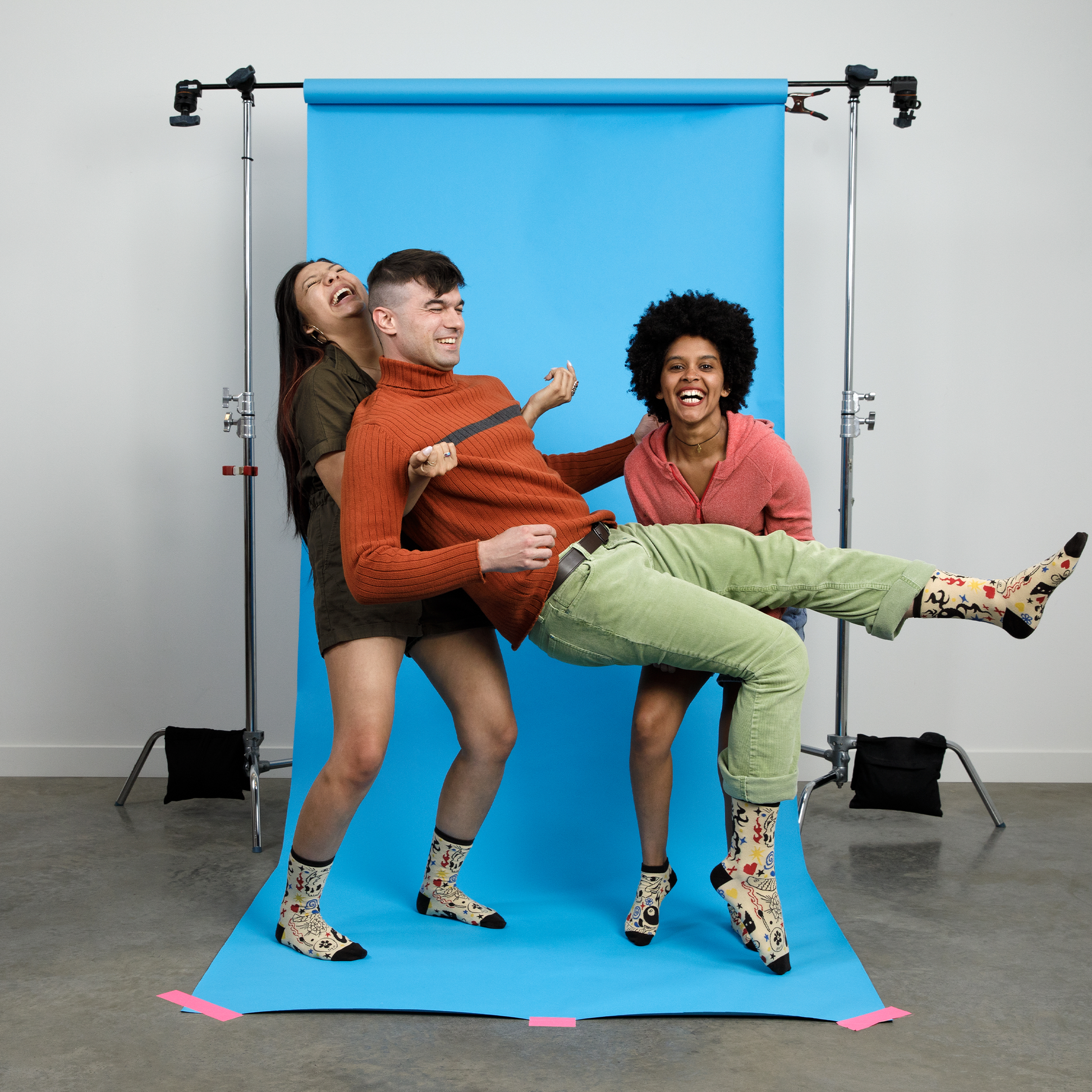 Three friends posing playfully against a bright blue photography backdrop. They're all wearing matching patterned Awesome Socks and laughing heartily. One person wears an orange turtleneck sweater and green pants, another wears a dark top, and the third wears a pink hoodie. They're arranged in a dynamic pose suggesting movement and joy. 