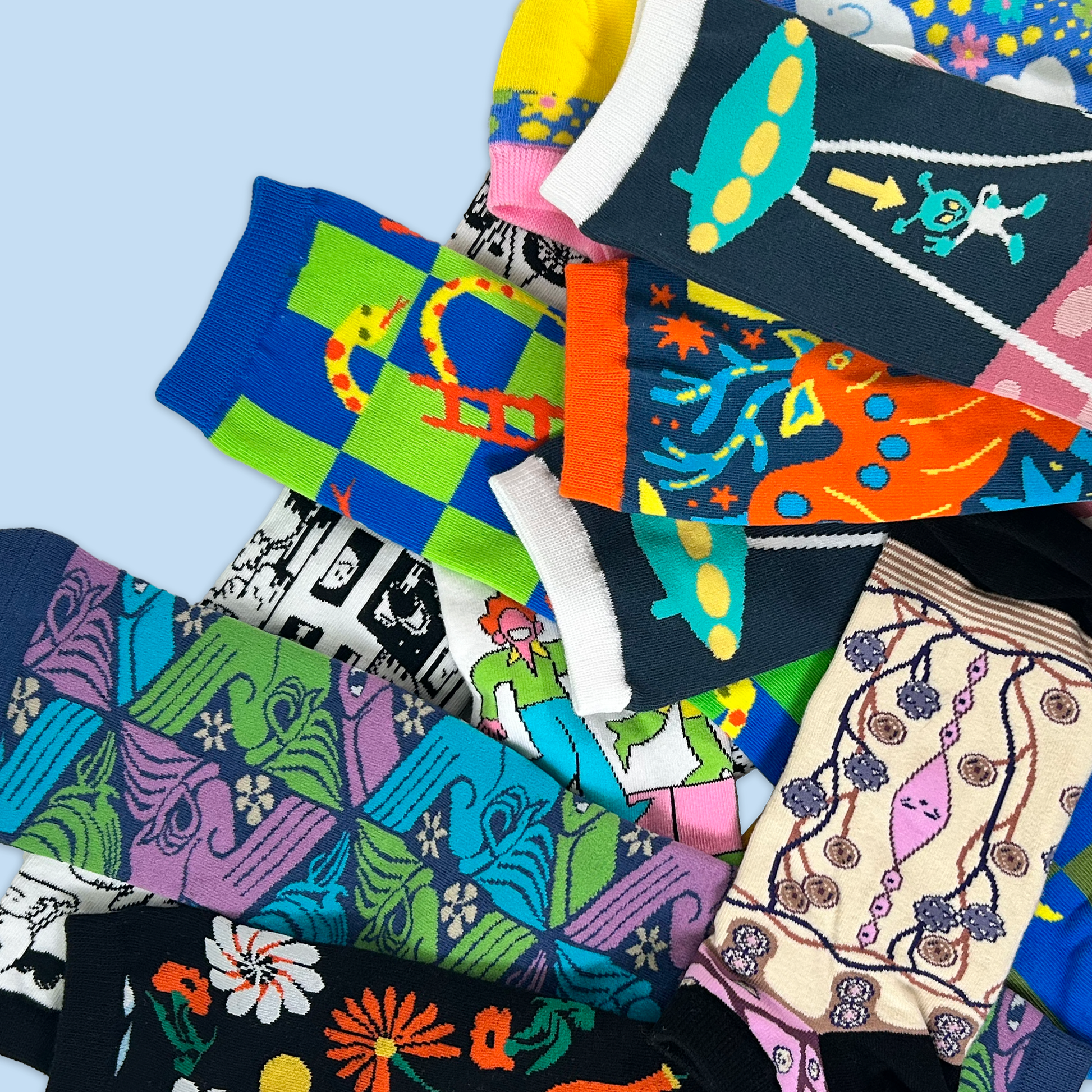 A collection of colorful patterned socks laid overlapping each other against a light blue background. The socks feature various designs including floral patterns, geometric shapes, abstract designs, and whimsical characters. The color palette includes bright blues, greens, purples, oranges, yellows, and pinks. Some socks have white trim and the patterns range from modern to traditional styles.