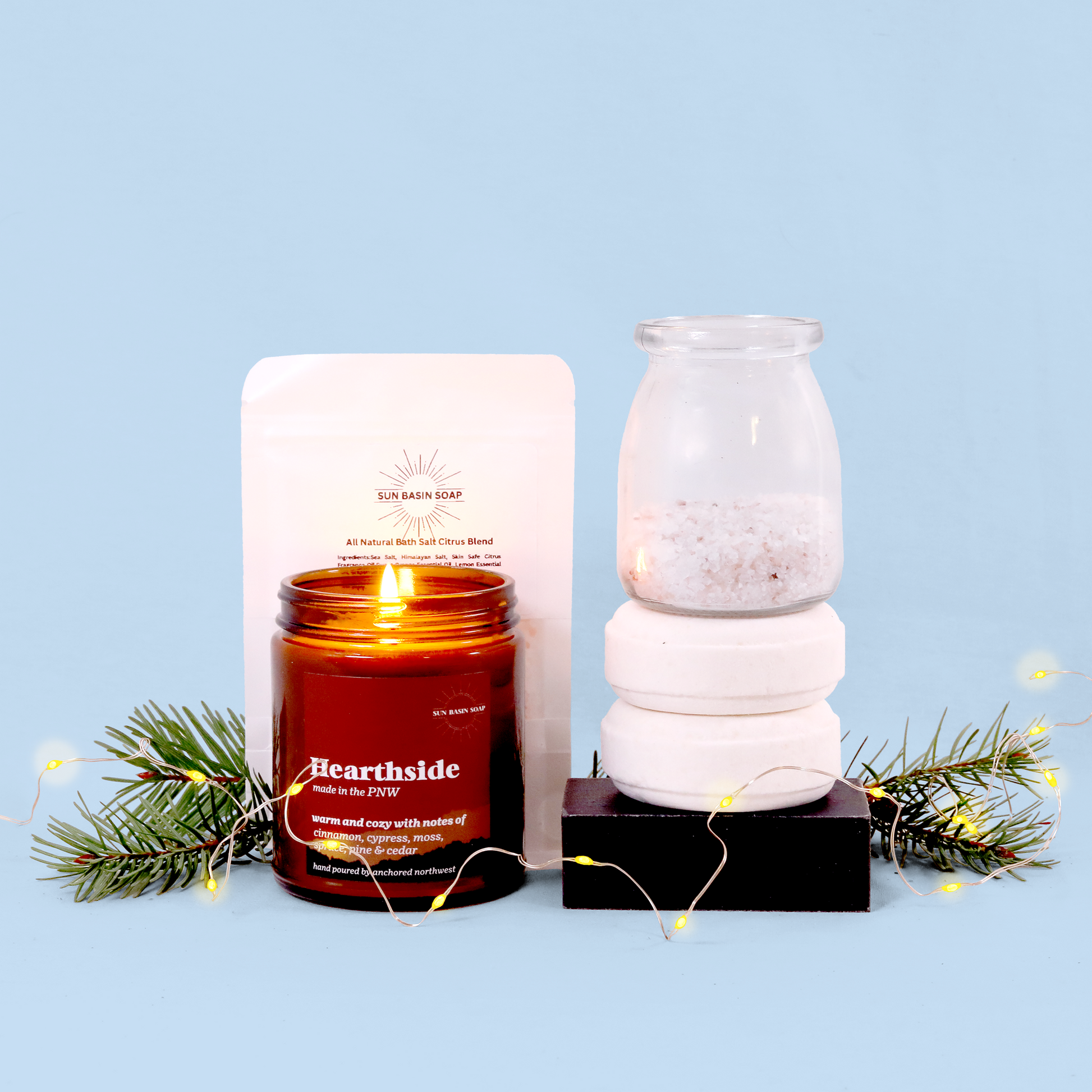 Product photo of self-care items against a light blue background. Features a lit amber-colored "Hearthside" candle by Sun Basin Soap with notes of cinnamon and cedar, alongside stacked bath products including what appears to be bath salts in a glass jar and white soap bars on a black platform. The scene is decorated with pine branches and small yellow fairy lights, creating a cozy winter atmosphere.