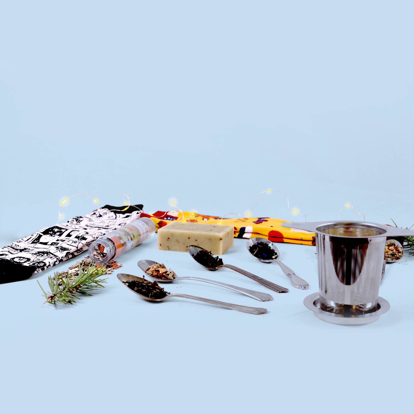 Wide-angle view of the same product arrangement showing additional elements including a silver tea strainer with base, loose tea samples on spoons, a natural soap bar, patterned socks, and a pine sprig with fairy lights, all arranged in a line against a light blue background.