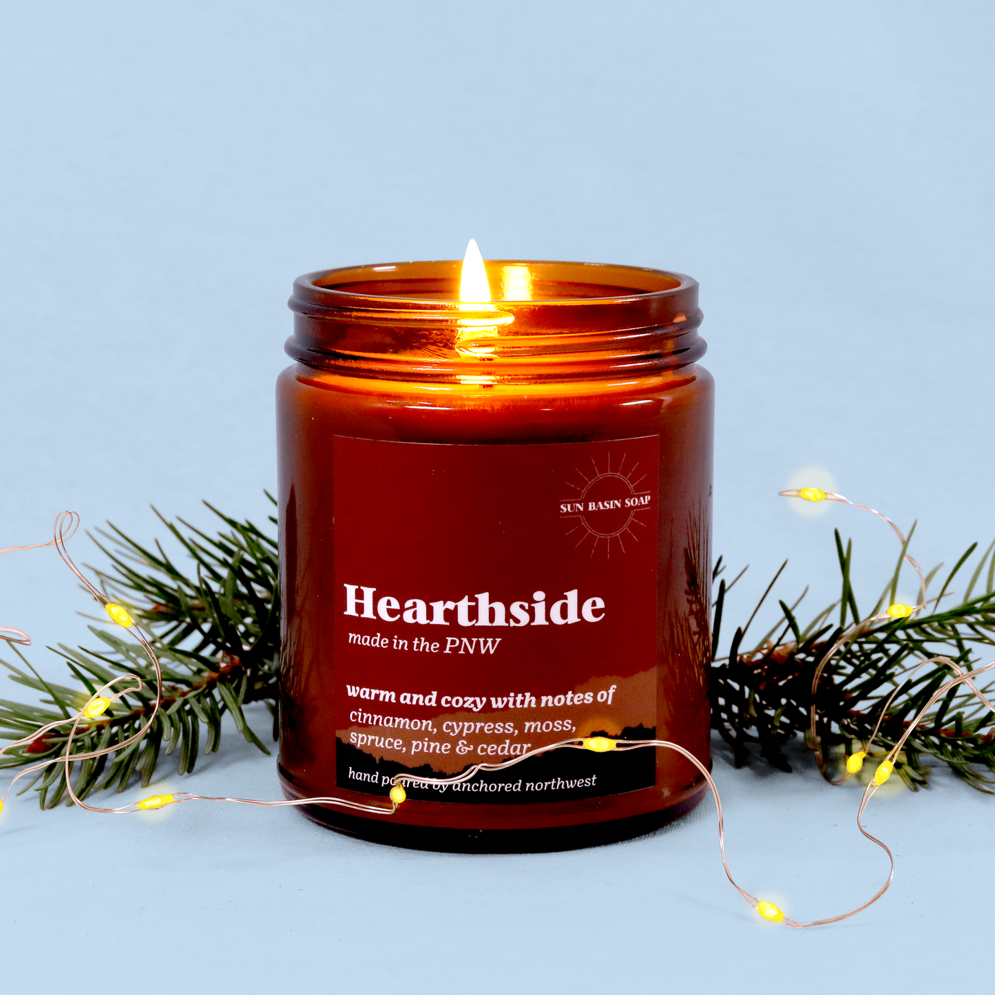 A lit amber glass candle labeled "Hearthside" by Sun Basin Soap, featuring notes of cinnamon, cypress, moss, spruce, pine and cedar. The candle is decorated with evergreen branches and delicate fairy lights against a light blue background, creating a warm and cozy winter atmosphere.