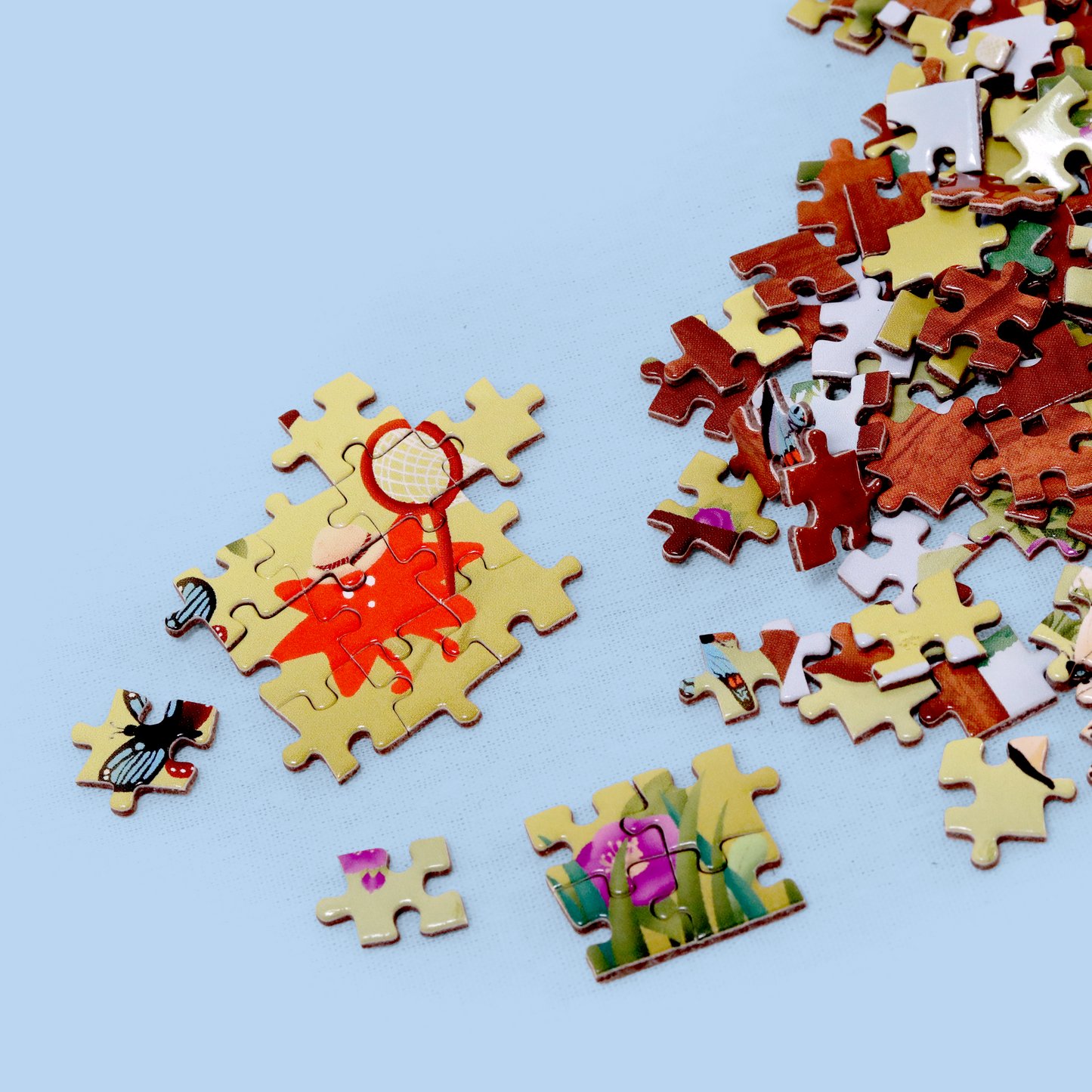 Scattered jigsaw puzzle pieces on a light blue background. A small cluster of connected pieces shows part of a red figure holding a magnifying glass. Other pieces display various colors and patterns, including flowers and insects.