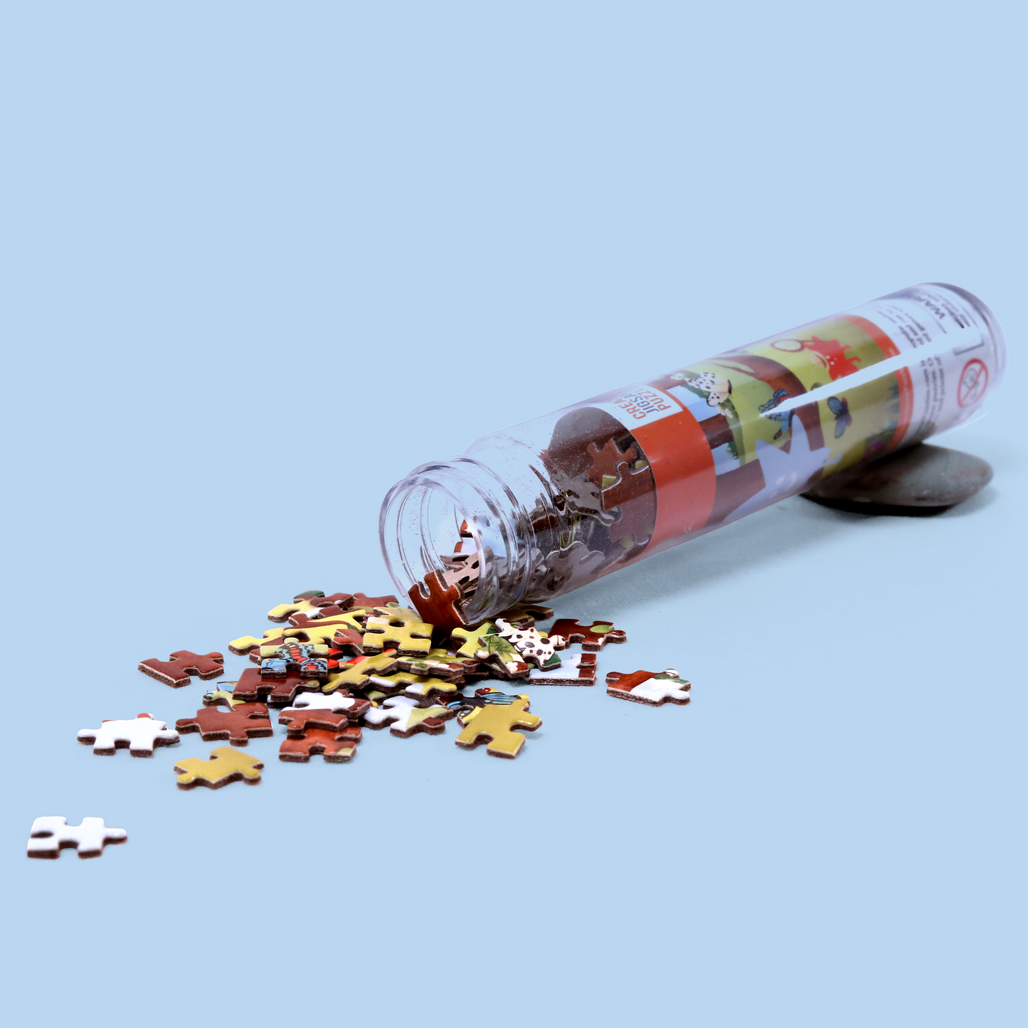 A clear plastic tube lying on its side with jigsaw puzzle pieces spilling out onto a light blue surface. The tube has a colorful label, and the puzzle pieces are in various shades of brown, yellow, and white.