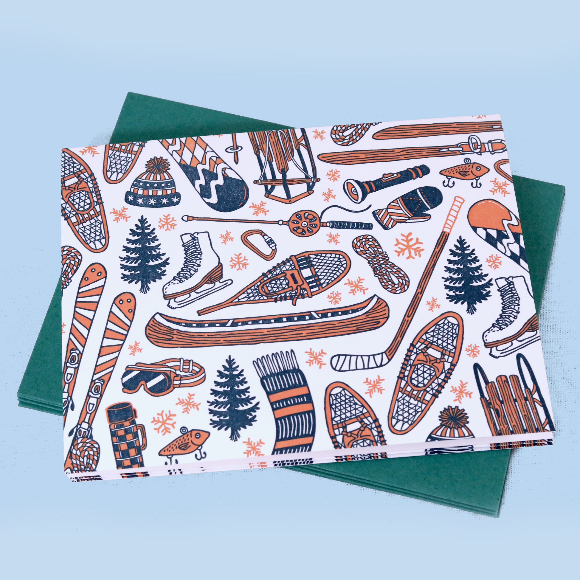 A winter-themed decorative card showing various outdoor activity items in navy blue and coral orange on white. The pattern includes snowshoes, skis, ice skates, hockey sticks, a canoe, winter hats, pine trees, and snowflakes, laid over a dark green envelope.