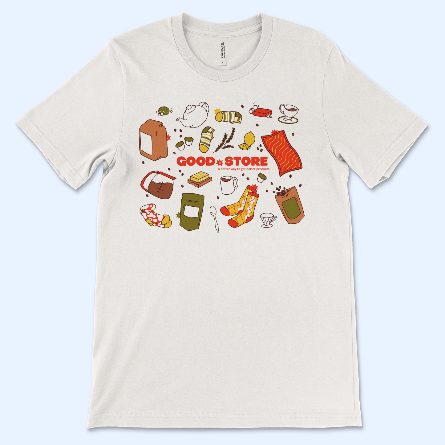 A close-up detail shot of the t-shirt design showing various illustrated food items including striped bread, wheat stalks, lemons, salmon fillet, coffee beans, and other grocery products scattered around the central "GOOD STORE" text in red.
