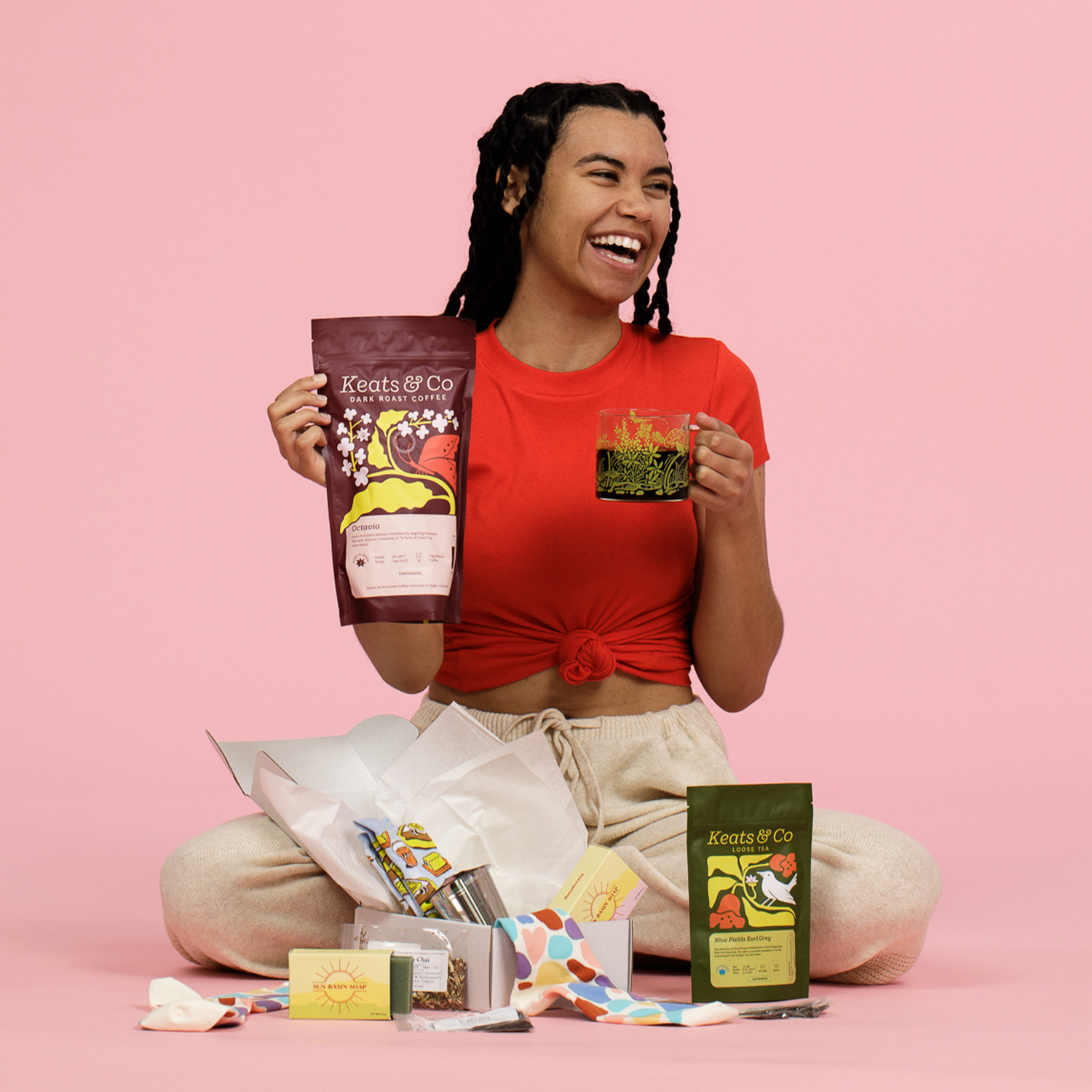 A lifestyle product photo showing someone wearing a red t-shirt and beige sweatpants, sitting cross-legged and laughing while holding Keats & Co coffee products. Various items are displayed in front of them including colorful socks, tea accessories, and both dark roast and loose tea packages.