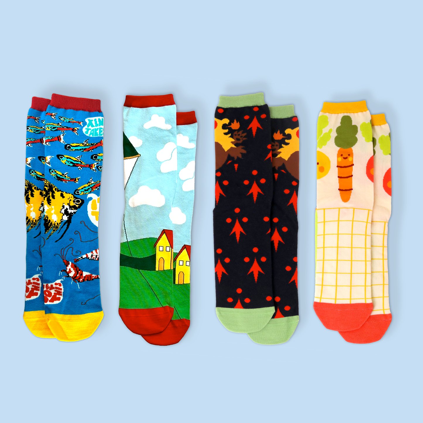 Four colorful patterned crew socks laid flat on a light blue background. From left to right: a blue sock with fish and wave patterns, a sock featuring a countryside scene with yellow houses and white clouds, a black sock with red leaf or arrow patterns, and a cream-colored sock with a cute carrot character and grid design. Each sock has a different colored cuff and heel.