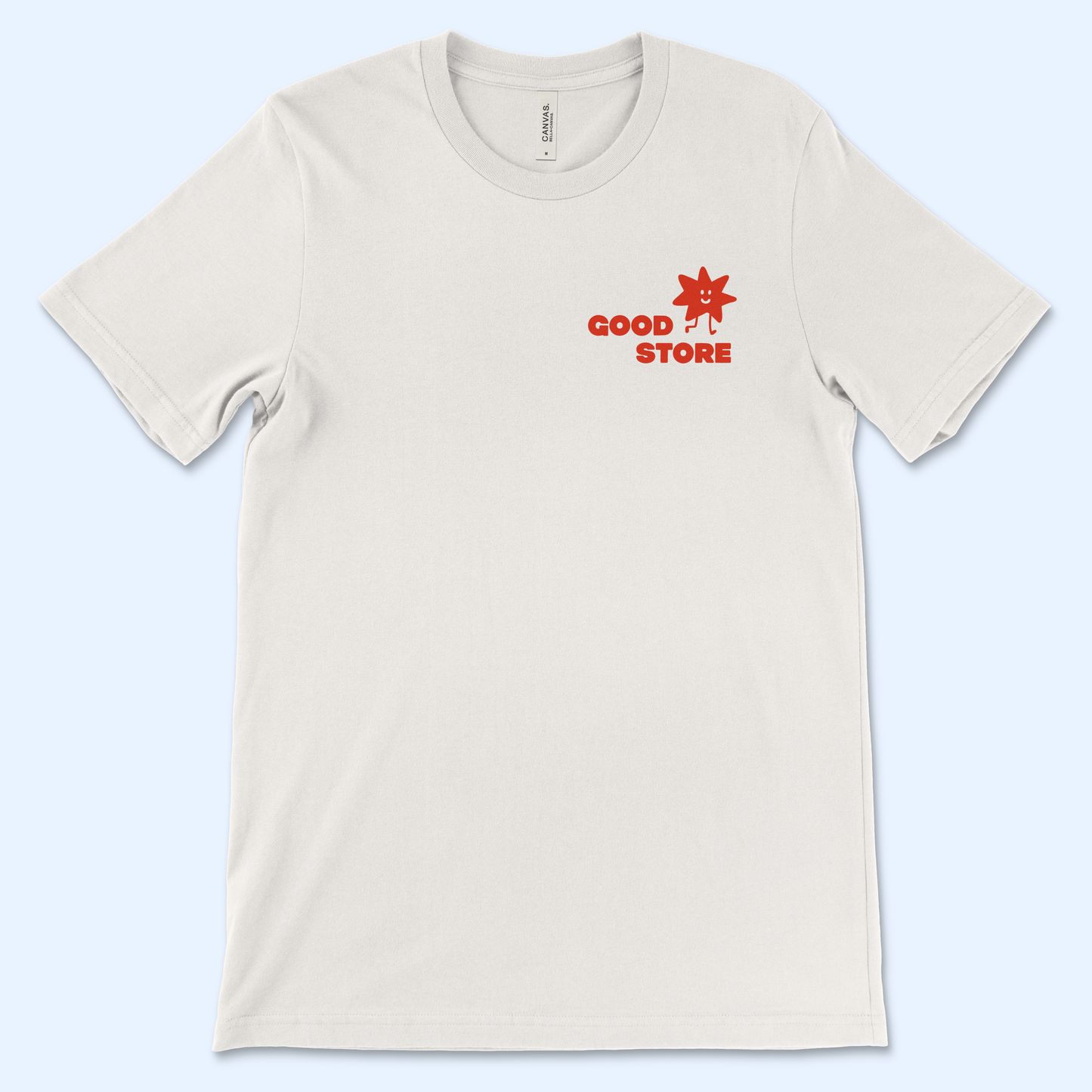 A single beige t-shirt on a light blue background featuring a small red "GOOD STORE" logo on the upper left chest area, accompanied by a cheerful red star mascot with a smiling face and stick arms.