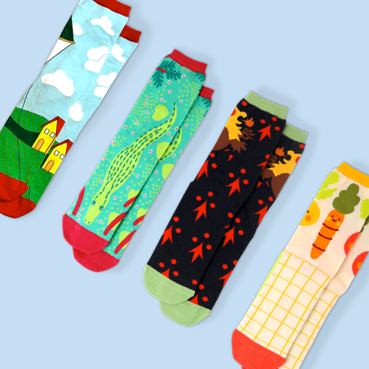 Four novelty socks arranged diagonally on a light blue background. From left to right: a sock with a countryside scene featuring yellow houses and white clouds, a turquoise sock with a whimsical alligator and flower pattern, a black sock with autumn leaves and red dots, and a cream-colored sock with a grid pattern and cartoon carrots. Each sock has contrasting heel and toe colors and is photographed flat.