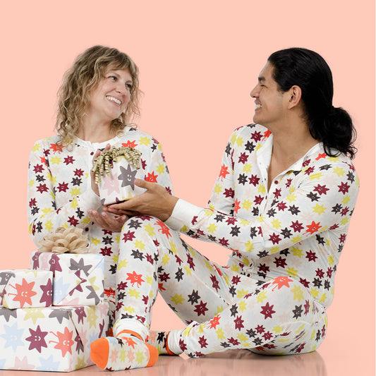 A smiling couple wearing matching white pajamas with a colorful floral star pattern sits together on a peach background, with wrapped gift boxes at their feet.
