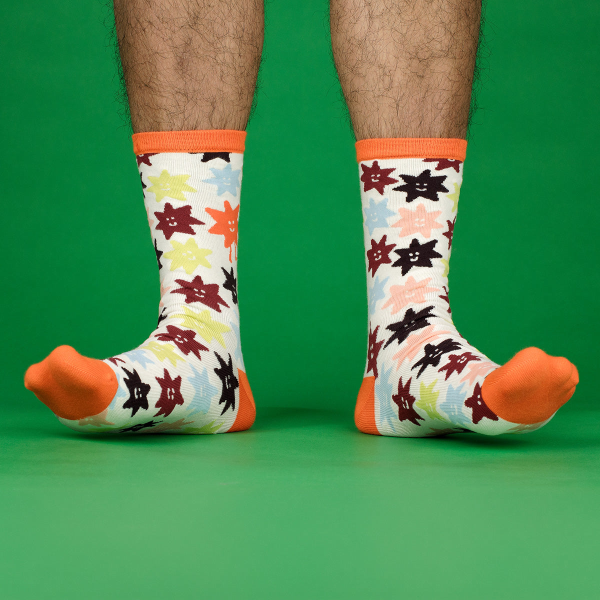 A pair of crew socks with an all-over colorful star pattern and bright orange trim, worn on a person's feet against a solid green background.