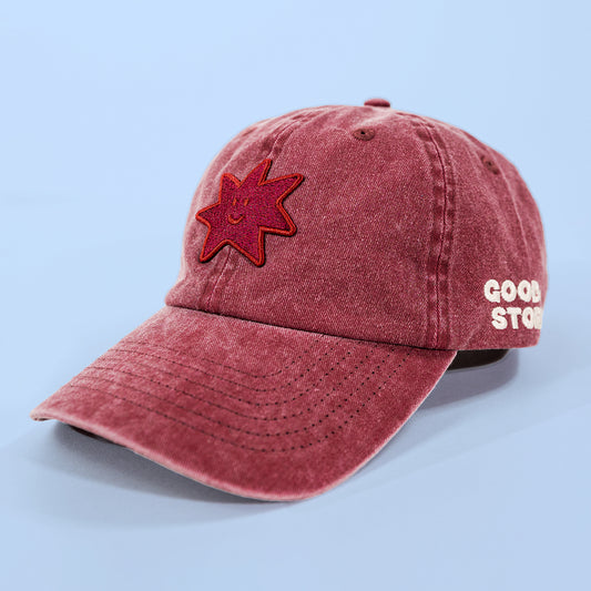A washed dusty rose baseball cap with a red embroidered star patch on the front and white embroidered text reading 'GOOD STORE' on the side, shown against a light blue background.