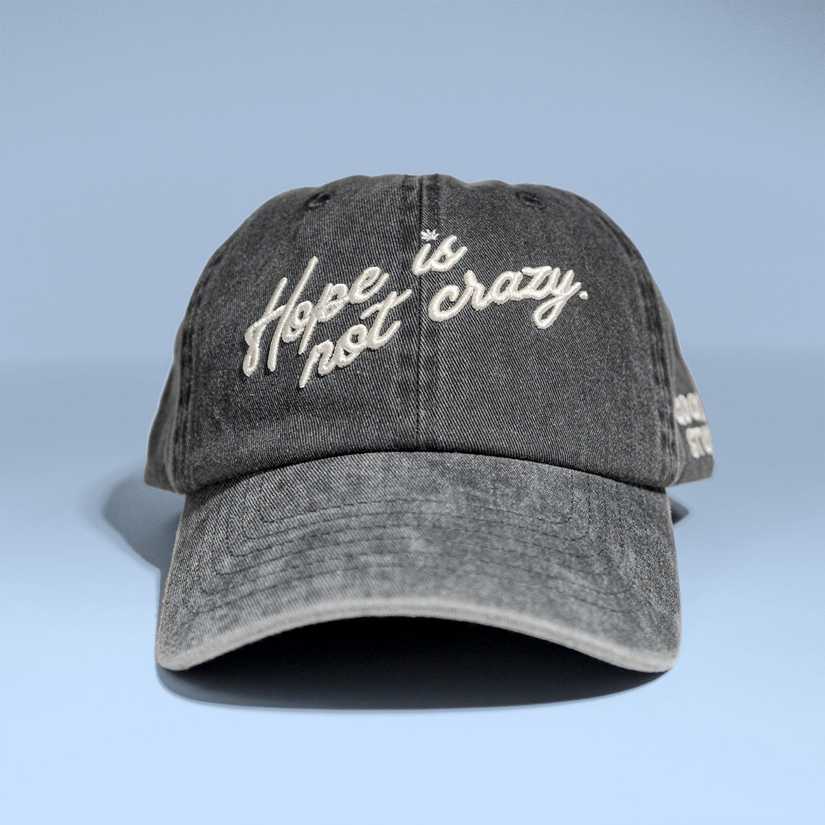 A dark gray acid-wash baseball cap with 'Hope is not crazy.' embroidered in white script across the front, set against a light blue gradient background.