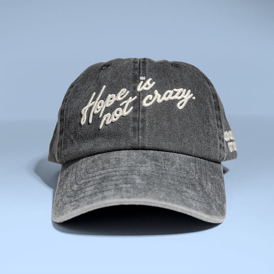 A dark gray acid-wash baseball cap with 'Hope is not crazy.' embroidered in white script across the front, set against a light blue gradient background.