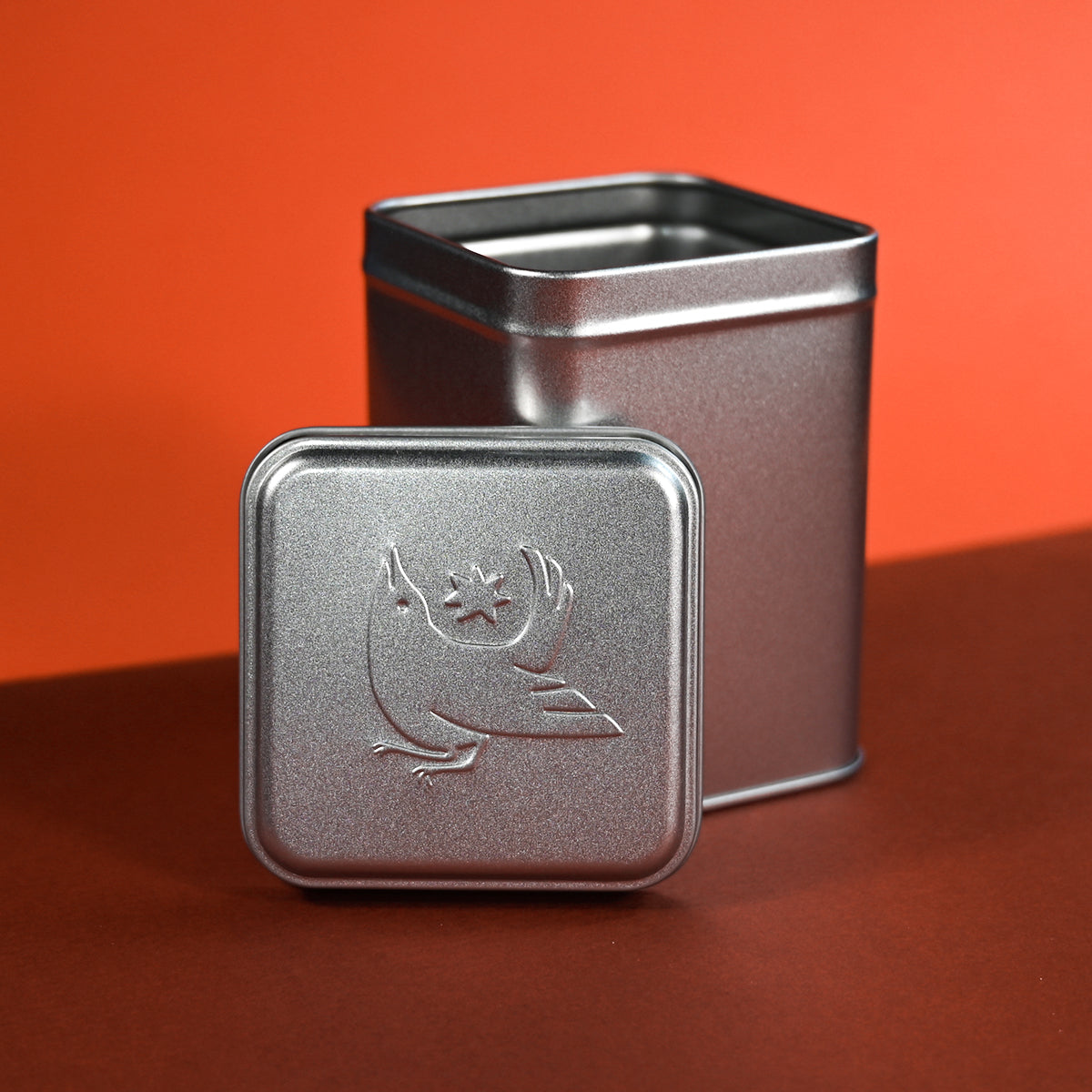 A square silver metal tin with its open top visible, standing behind its matching lid. The lid is propped upright in front and features a embossed illustration of a bird with its head tilted up toward a small star, with wings raised and tail feathers fanned. The tins sit on a dark red-brown surface against a vivid orange-red backdrop.