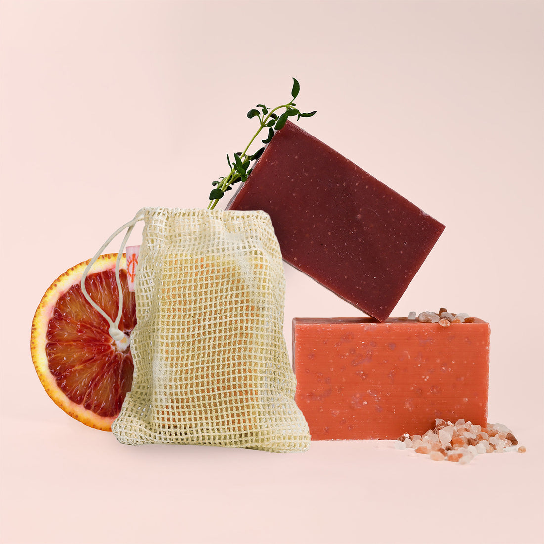 Three bars of soap with a mesh bag and a slice of blood orange on a light pink background