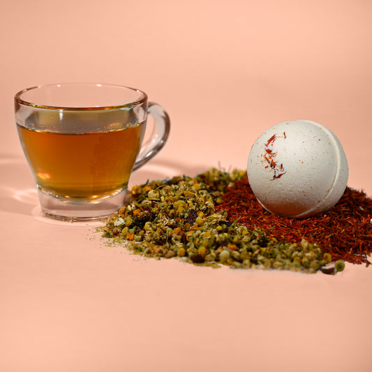 Clear glass mug with tea, dried herbs, and a bath bomb on a pink background