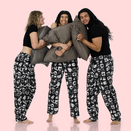Three people standing barefoot against a solid pale pink background, wearing matching black pajama tops and black pants with a white cartoon-style graphic print. The person in the center holds two gray pillows.