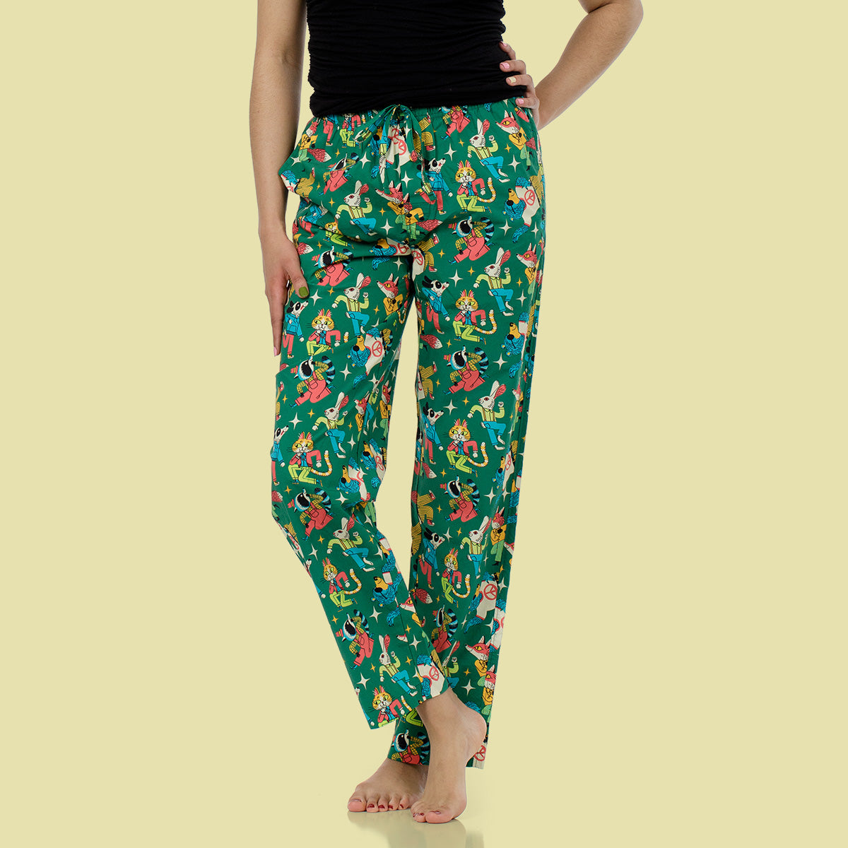 A person modeling green drawstring pants with a colorful cartoon-style print, paired with a black top, against a pale yellow background.