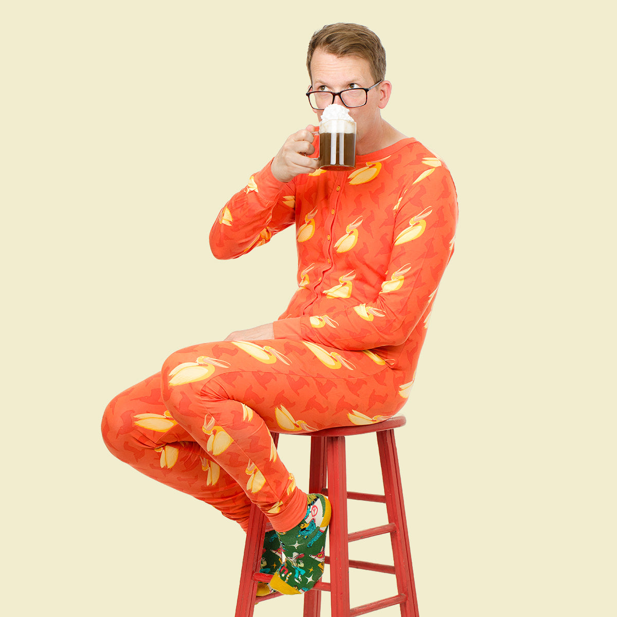 A man wearing a bright orange one-piece pajama suit with a yellow pelicans pattern sits on a red stool, holding a mug to his mouth against a solid pale yellow background.