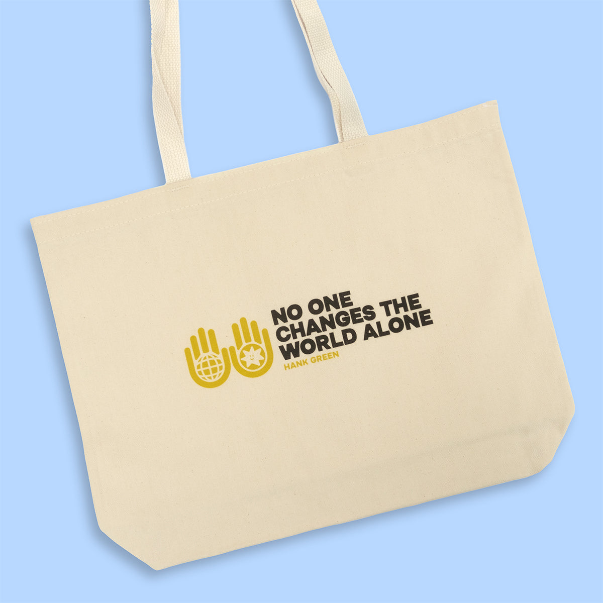 Natural canvas tote bag with cream-colored handles on a light blue background, featuring two yellow hand icons holding globes alongside dark brown text reading 'NO ONE CHANGES THE WORLD ALONE' attributed to Hank Green