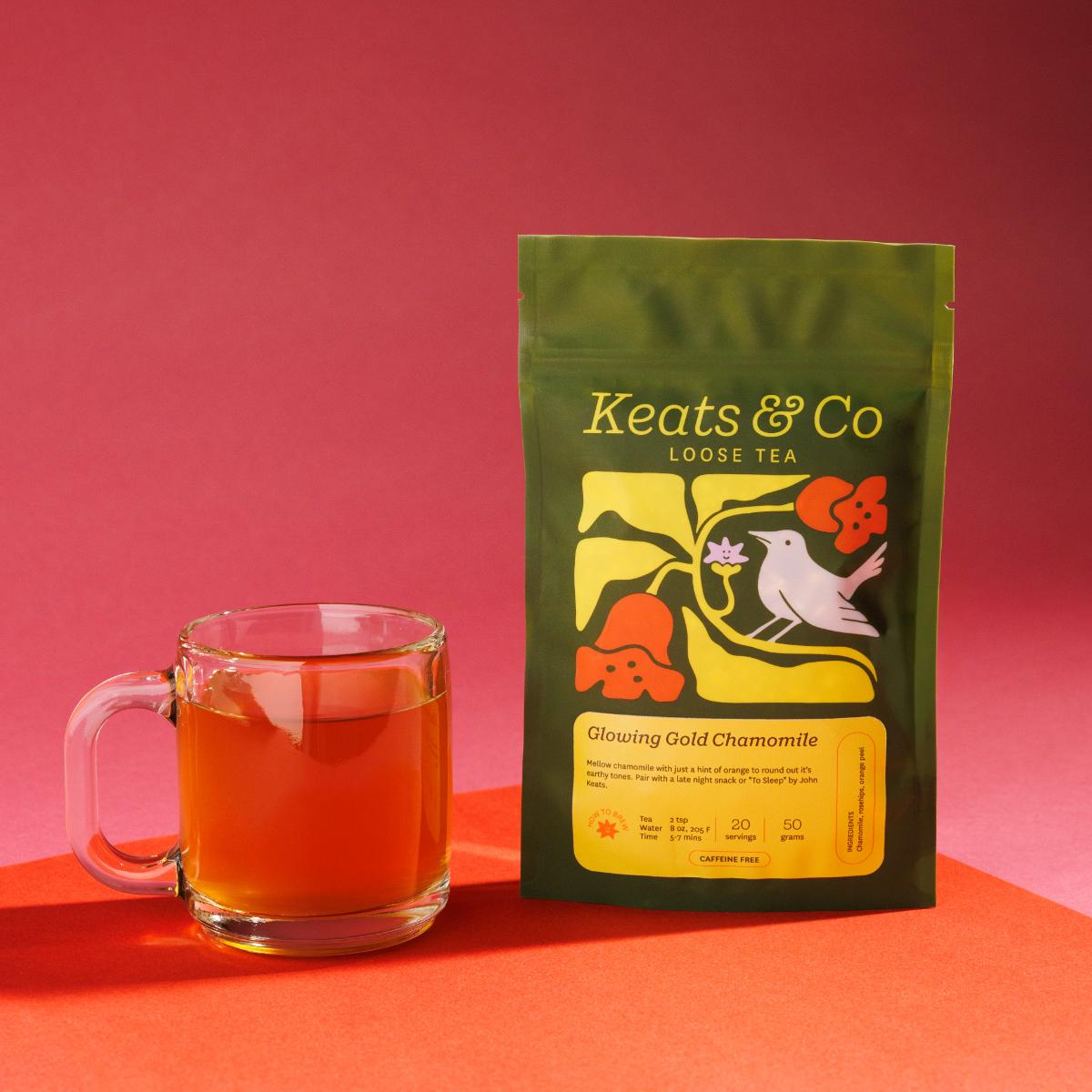 Green pouch of Keats & Co Loose Tea Glowing Gold Chamomile against a pink background, featuring illustrated artwork of a white bird and orange flowers, displayed next to a clear glass mug filled with an orange brewed tea on an orange surface.