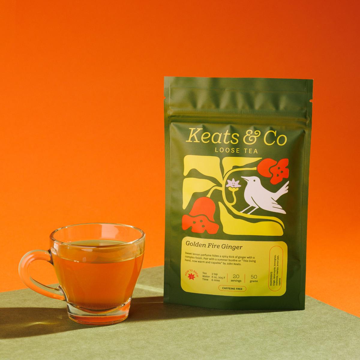 Green pouch of Keats & Co Loose Tea Golden Fire Ginger against an orange background, featuring illustrated artwork of a white bird and orange flowers, displayed next to a clear glass mug filled with yellowish brewed tea on a green surface.