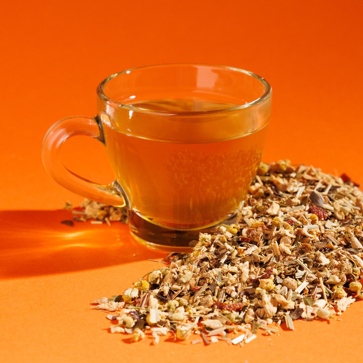 Glass cup of orange Golden Fire Ginger tea positioned next to a pile of loose leaf herbals on an orange background. A Good Store product.
