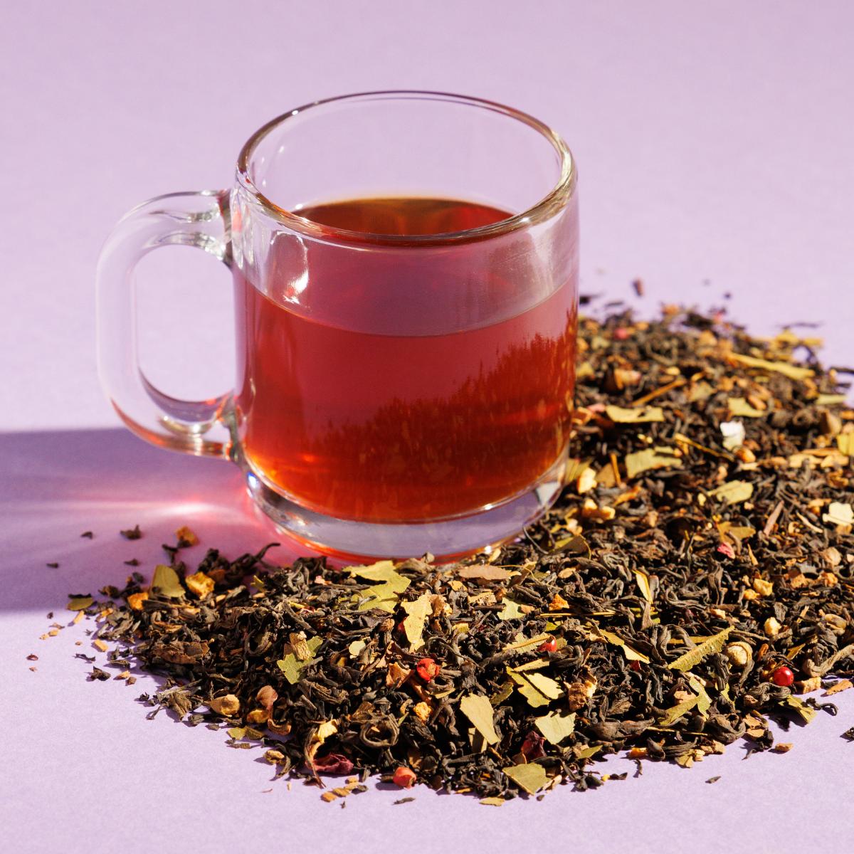 Glass cup of redish Golden Moon tea positioned next to a pile of loose leaf tea blend containing on a purple background. A Good Store product.