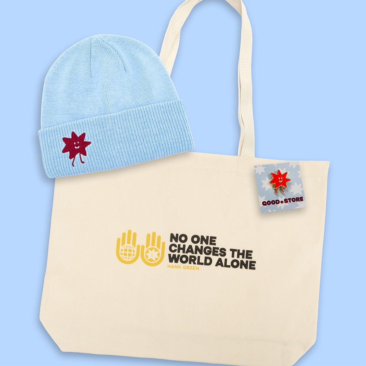 Light blue beanie with red star design, beige tote bag with text and logo, and small enamel pin on a light blue background