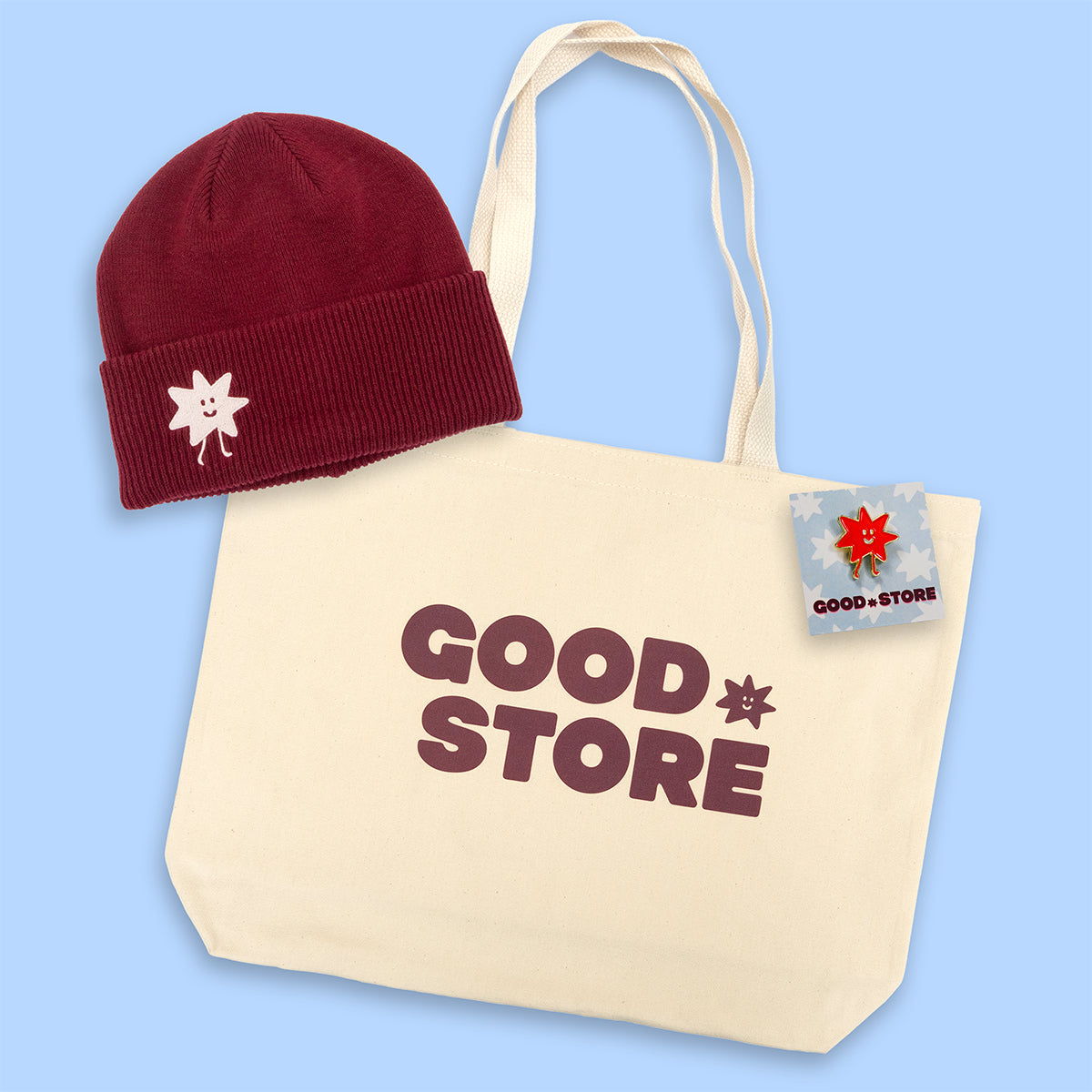 Beige tote bag with 'GOOD STORE' text, maroon beanie with white logo, and small enamel pin on a light blue background