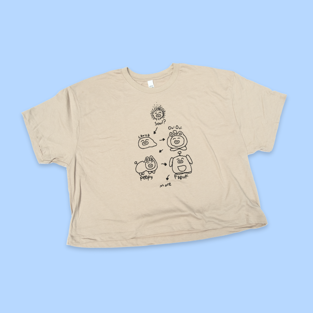 A tan crop top laid flat on a light blue background. The shirt features a black line drawing showing the "evolution" or life cycle of a cute character, with arrows connecting different stages: starting with "Soul" (a spiky circular design), then "Larva" (a simple blob shape), "Ow Ow" (a round character with a cap), "Puppy" (a dog-like figure), and "Papaw" (an elderly character). The design has a playful, hand-drawn aesthetic with simple cartoon-style illustrations.