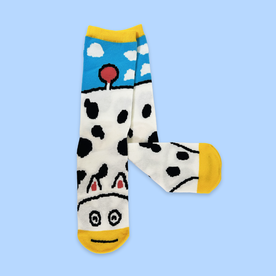 A pair of white socks with black cow spots and yellow toe/heel accents. The top features a blue sky section with white clouds. The bottom of each sock has a cartoon cow face with black and white eyes and a small black smile.