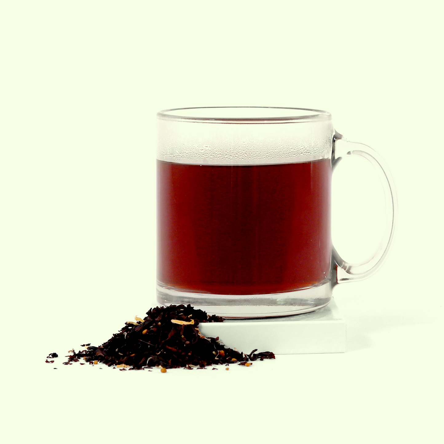 A clear glass mug containing brewed dark reddish-brown tea. In front of the mug is a small pile of loose black tea leaves mixed with lighter colored ingredients.