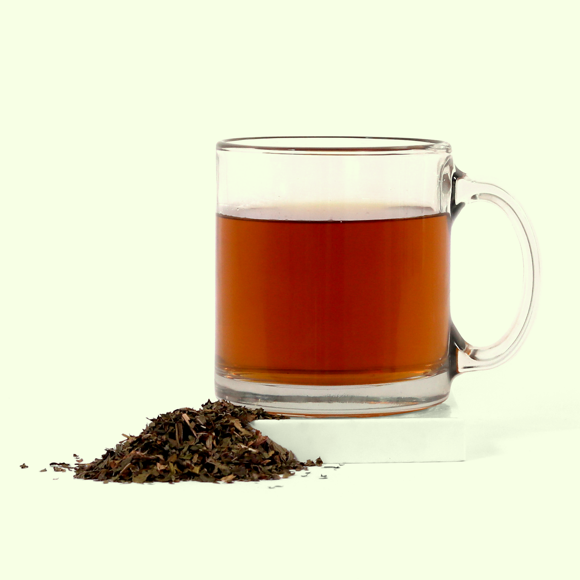 A clear glass mug filled with brewed amber-colored tea. In front of the mug is a small pile of loose dried tea leaves and herbs in various shades of brown and green against a white background.