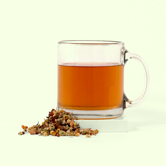 A clear glass mug containing amber-colored tea. Next to the mug is a small pile of loose herbal tea mixture consisting of dried flowers, herbs, and small buds in various shades of brown, beige, and orange.