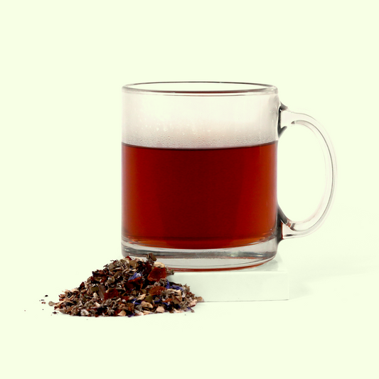 A clear glass mug containing brewed red tea. Next to the mug is a small pile of loose herbal tea blend containing various dried herbs, flower petals, and plant materials.