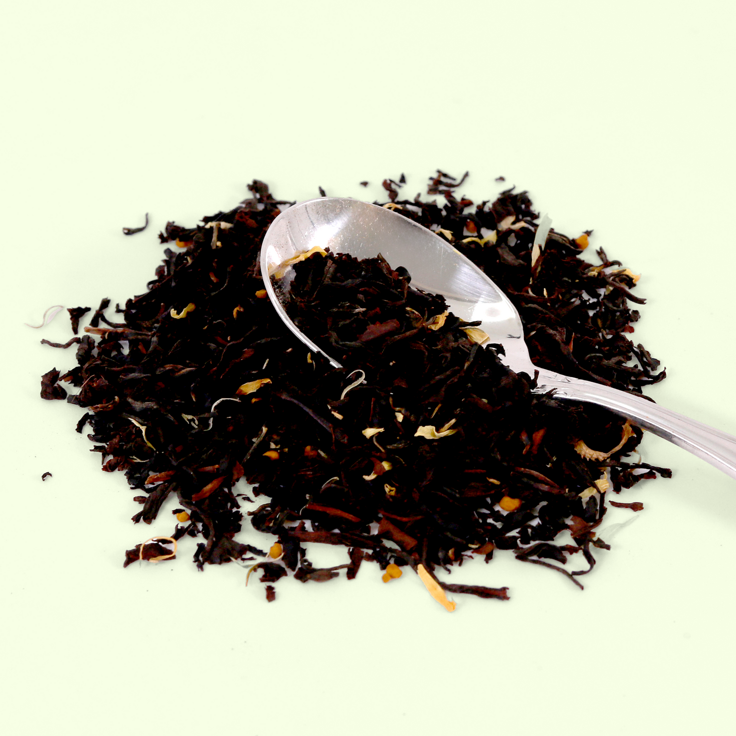 A close-up of loose black tea leaves with scattered lighter colored bits (possibly flower petals, citrus peel, or spices) arranged in a pile with a silver measuring spoon resting on top. The tea mixture is displayed against a pale background.