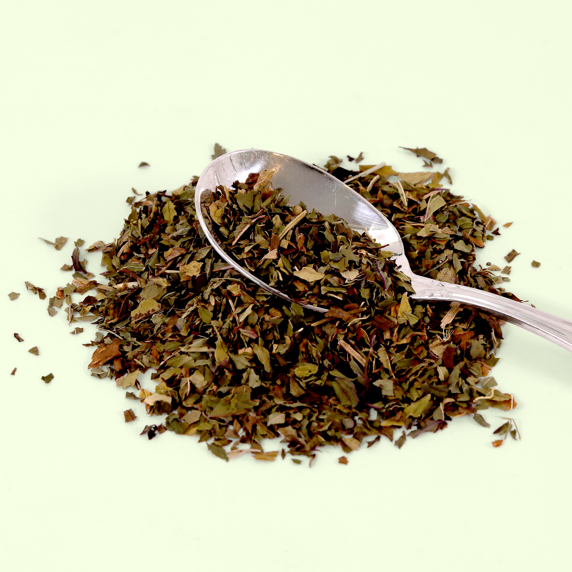 A close-up of loose dried herbs and tea leaves in shades of green, brown, and red with a silver measuring spoon resting on top. The mixture appears to be primarily composed of dried leaves and small stems displayed against a pale background.