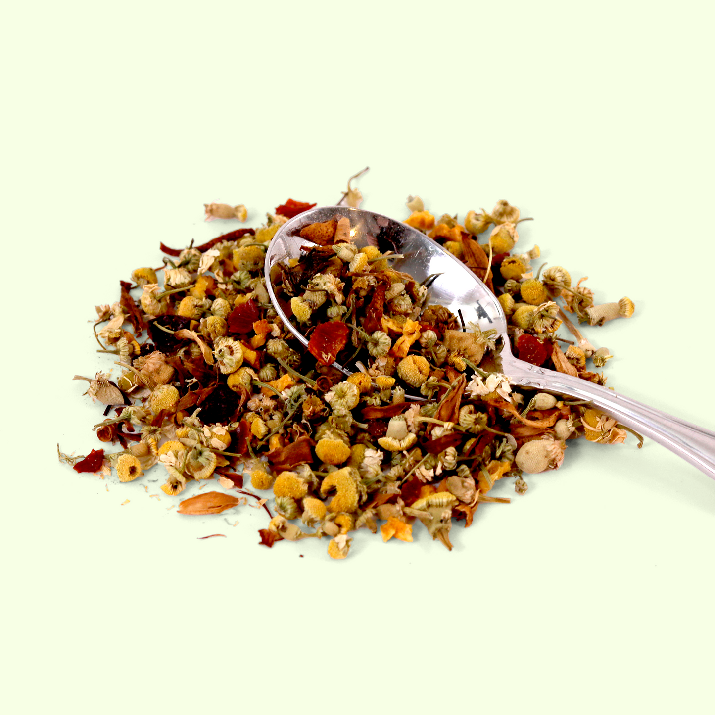 A close-up of a loose herbal tea blend with a silver measuring spoon placed in the mixture. The blend contains predominantly light-colored ingredients including dried chamomile flowers (small yellow buds), orange petals, and various other dried herbs and flowers in shades of beige, yellow, and red.