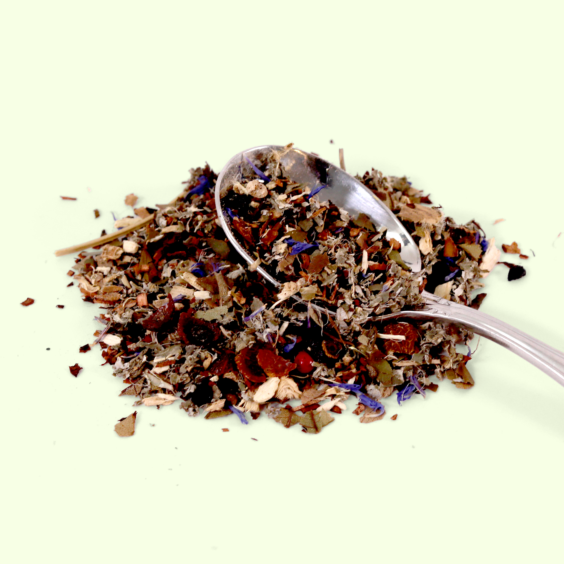 A close-up of the loose herbal tea blend with a silver measuring spoon resting in it. The blend consists of various dried plant materials including what appears to be bark pieces, flower petals (some blue/purple), dried berries or fruits, and herbs of different textures and colors.