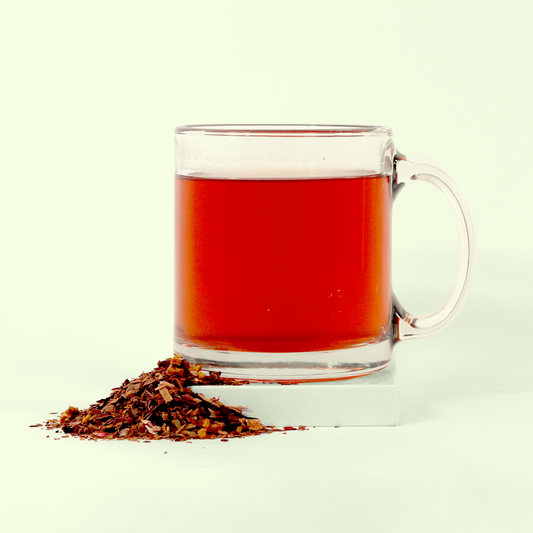 A clear glass mug filled with reddish-brown tea next to a small pile of loose-leaf tea blend on a pale background. The tea appears to be brewed and has a rich amber-reddish color, while the dry tea leaves beside it show various flecks of different ingredients including what appear to be pieces of fruit or herbs mixed with the tea leaves.