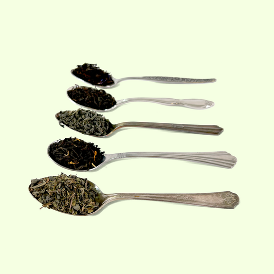Five ornate silver spoons arranged vertically, each containing different varieties of loose leaf tea. From top to bottom: dark black tea leaves, medium black tea, green tea leaves, black tea with visible petals or additions, and a lighter green tea blend. The spoons have decorative handles with various vintage patterns.