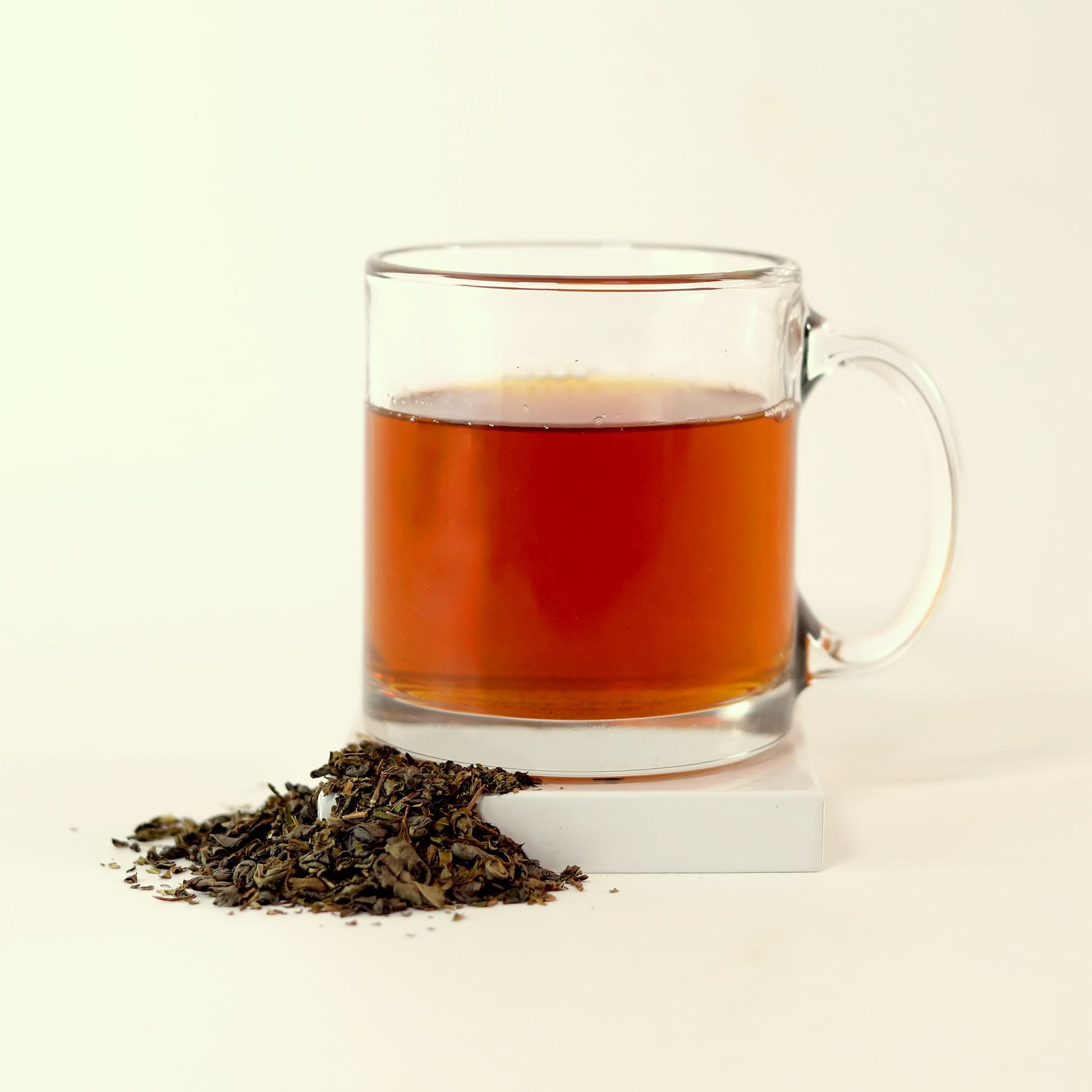  A clear glass mug filled with light orange tea - April Meadows Green -  next to a small pile of dried tea leaves. A Good Store product.