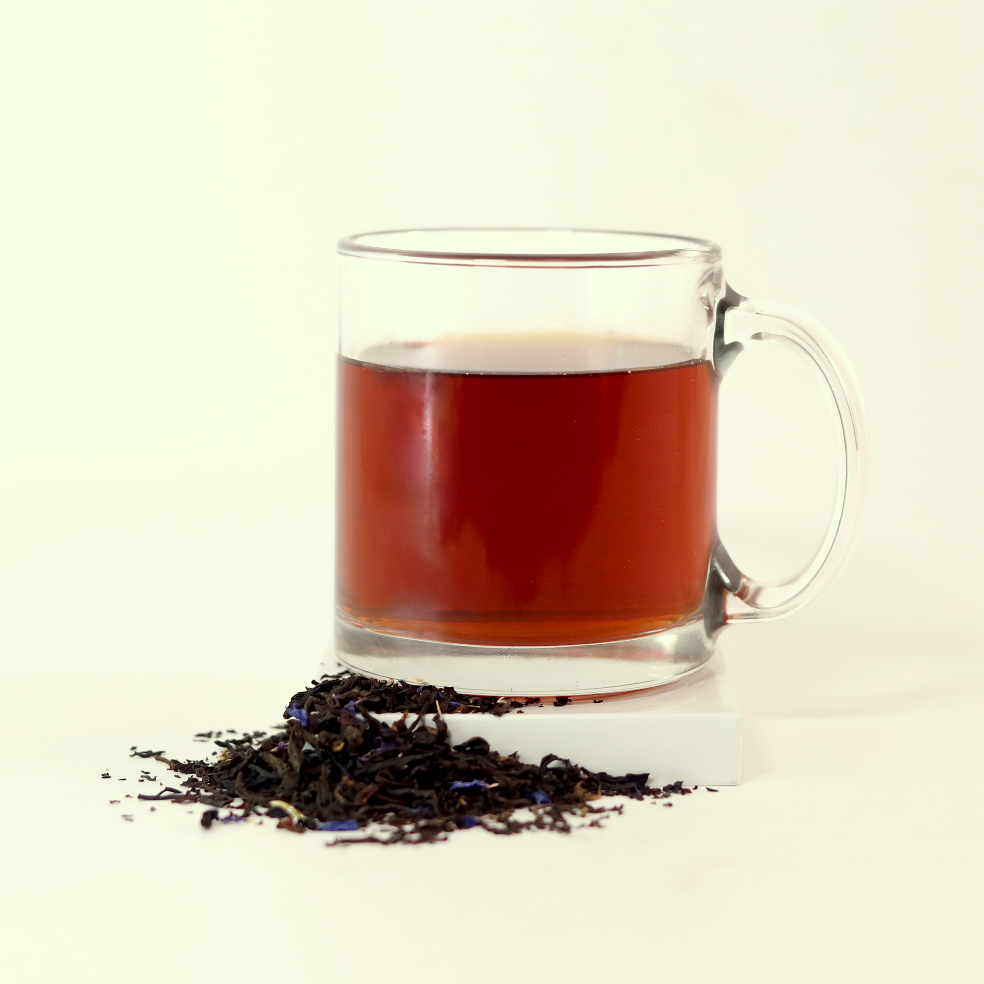  A clear glass mug filled with reddish-orange tea - Blue Fields Earl Grey -  next to a small pile of dried tea leaves. A Good Store product.
