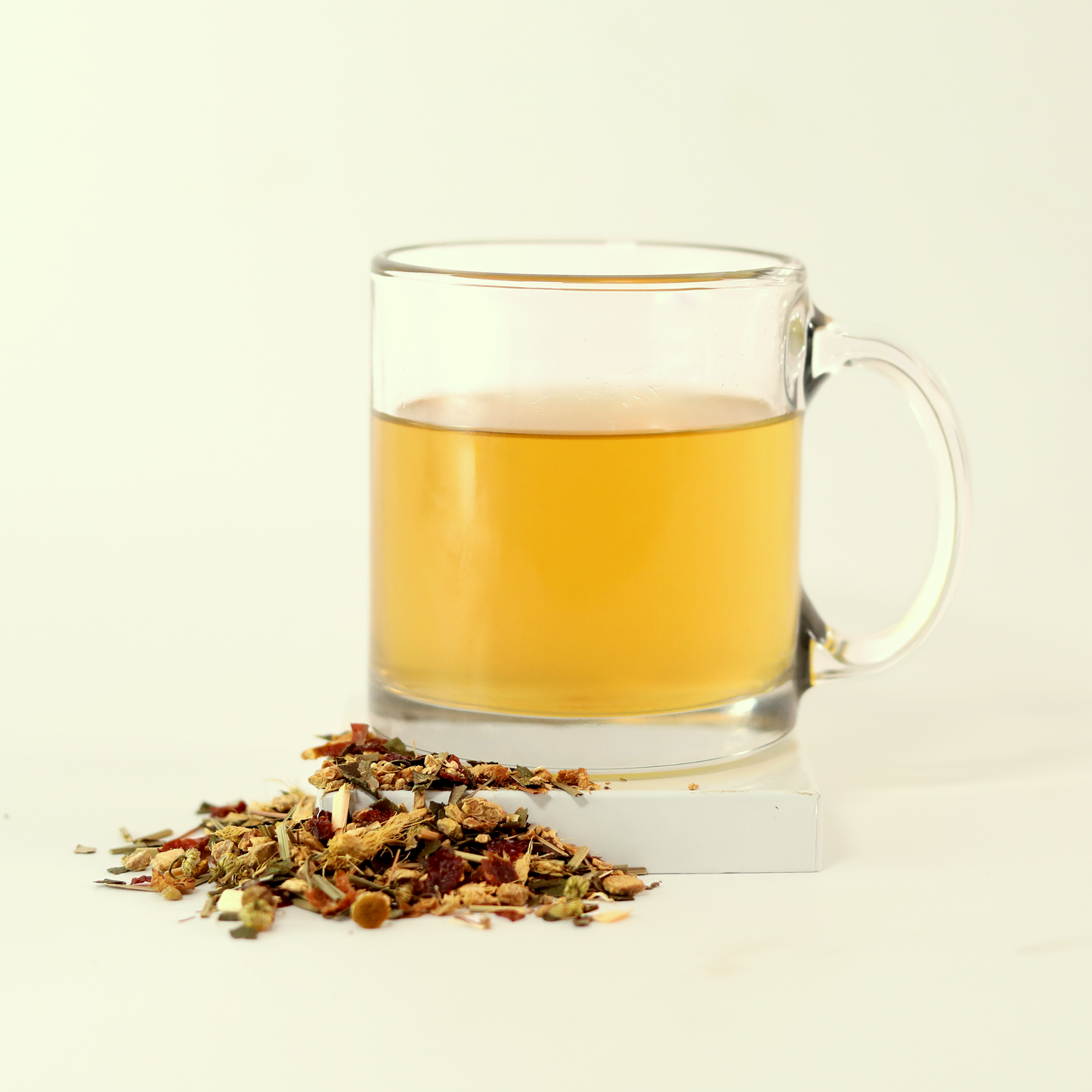  A clear glass mug filled with light yellow tea - Goldenfire Ginger -  next to a small pile of dried tea leaves. A Good Store product.